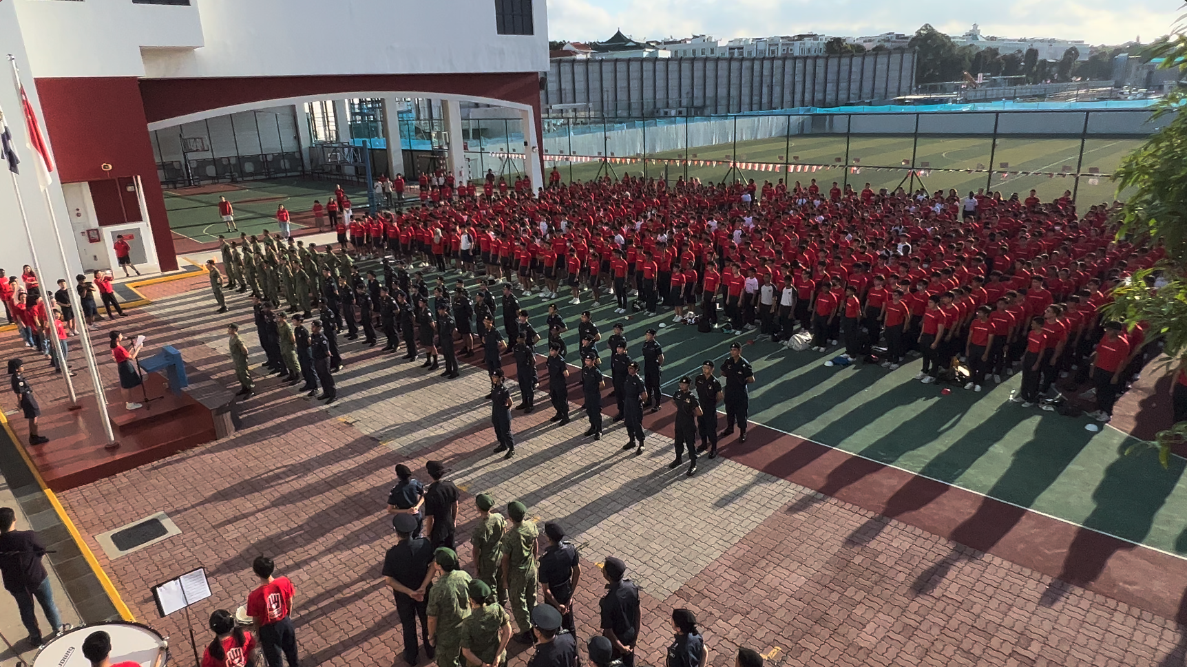 Large school assembly with hundreds of students in red shirts and uniformed cadets standing in formation outdoors.